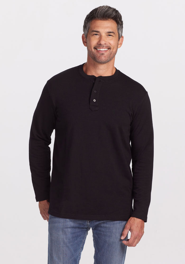 A man with short, dark graying hair and a beard is wearing the Jackson Henley - Black by Woolx, made of Merino wool, along with blue jeans. He is smiling and standing against a plain white background. 