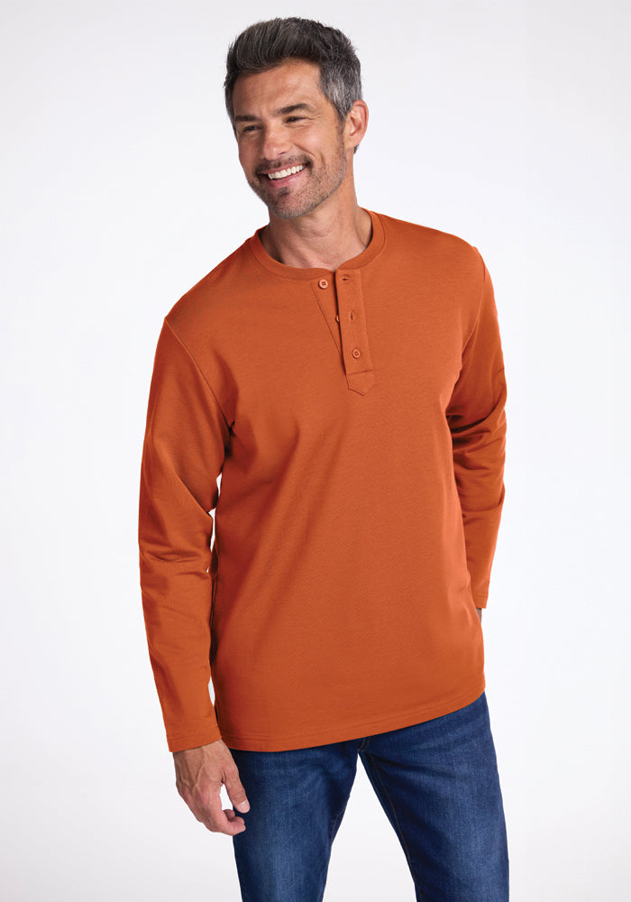 A man with short, gray hair and a beard smiles while wearing an orange Jackson Henley from Woolx paired with blue jeans. He stands against a plain, light-colored background with one hand in his pocket and the other at his side. 