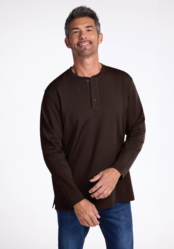 A man with short gray hair and a beard, wearing a dark brown Woolx Jackson Henley shirt and blue jeans, stands against a plain white background, smiling and resting his right hand on his left wrist. 