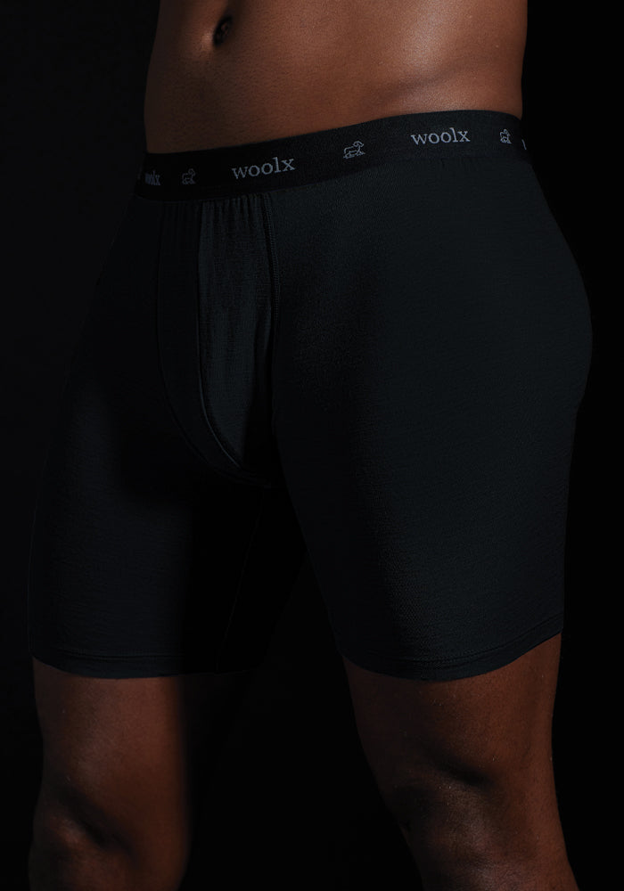 Against a dark backdrop, an individual models the black Jazzy No Fly Boxer Briefs by Woolx, showcasing their sleek fit and design with the signature "Woolx" waistband.