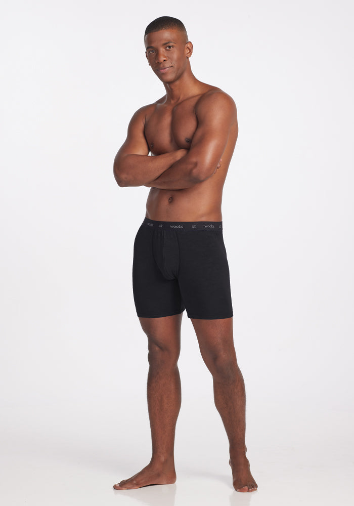 A man stands confidently against a plain white background, wearing black Jazzy No Fly Boxer Briefs by Woolx. He is barefoot and has his arms crossed, with a relaxed expression. The lightweight underwear features a subtly branded waistband, offering both style and comfort.