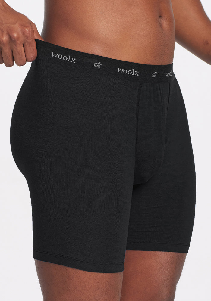 A person is wearing black Woolx Jazzy No Fly Boxer Briefs, visible from the waist to mid-thigh. The waistband prominently features the Woolx logo. The lightweight wool fabric looks smooth and fits snugly against the skin. The plain white background highlights the sleek design of these Merino boxer briefs.