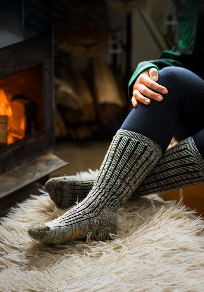 A person wearing the cozy and warm Joey Socks in Dark Forest Marled from Woolx sits cross-legged on a fluffy rug by a lit fireplace, with stacked firewood visible in the background.