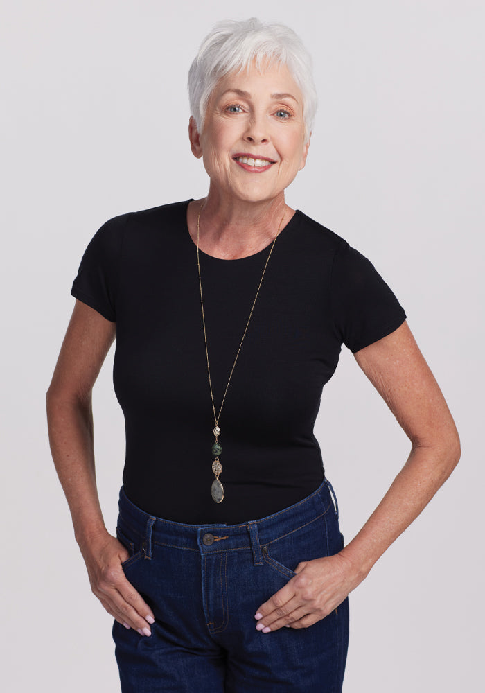 An older woman with short white hair smiles confidently, wearing the Woolx Gracie Bodysuit - Black, blue jeans, and a long necklace with a pendant. She stands with her hands in her pockets against a plain grey background. 