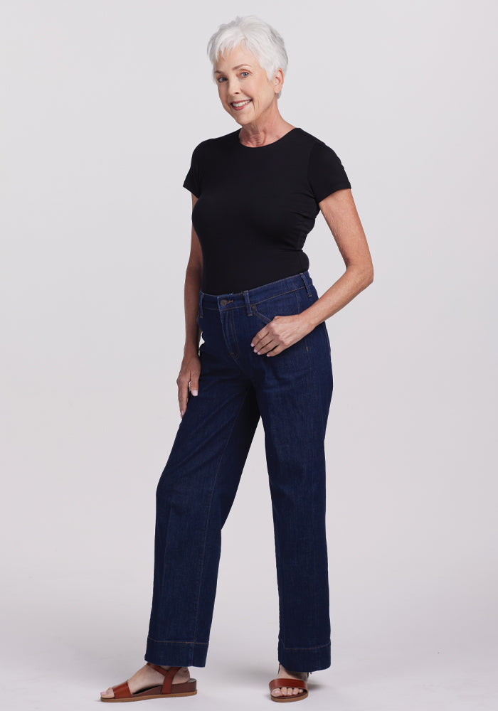 An older adult with short white hair is wearing a black Woolx Gracie Bodysuit, dark blue jeans, and brown sandals. They are smiling confidently against a plain white background.