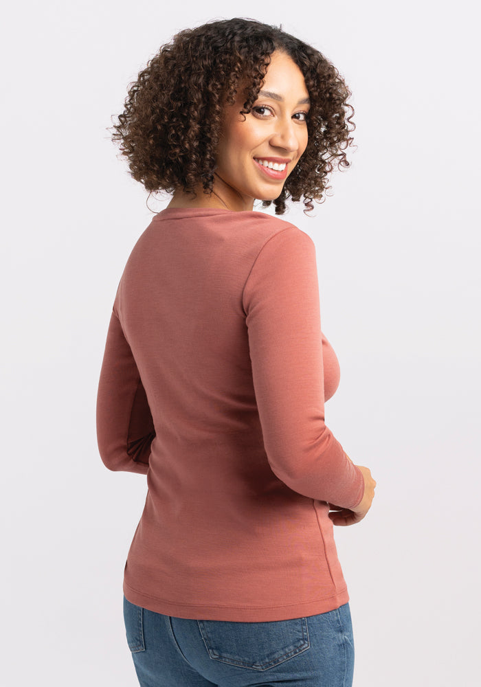 A woman with curly hair is smiling and looking back over her shoulder. She is wearing a long-sleeved, rose-colored Kenzie Scoop Neck Merino wool top from Woolx and blue jeans, perfect for business casual attire. The background is plain white.