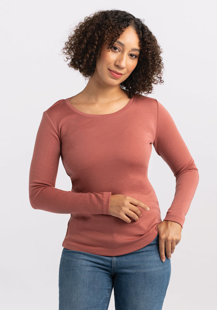 A person with curly hair is posing against a plain background, wearing a business casual, long-sleeved light red Kenzie Scoop Neck Merino wool shirt by Woolx and blue jeans. They have a neutral expression and are looking directly at the camera. One hand rests on their hip while the other hand is placed in front. 