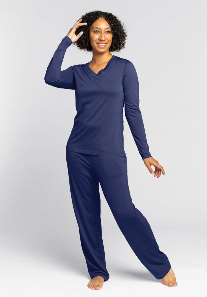 A person with curly hair smiles while wearing a matching Starry Night Lily V Neck Lounge Top by Woolx and pants. They stand barefoot on a light gray background, posing with one hand touching their hair and the other by their side.