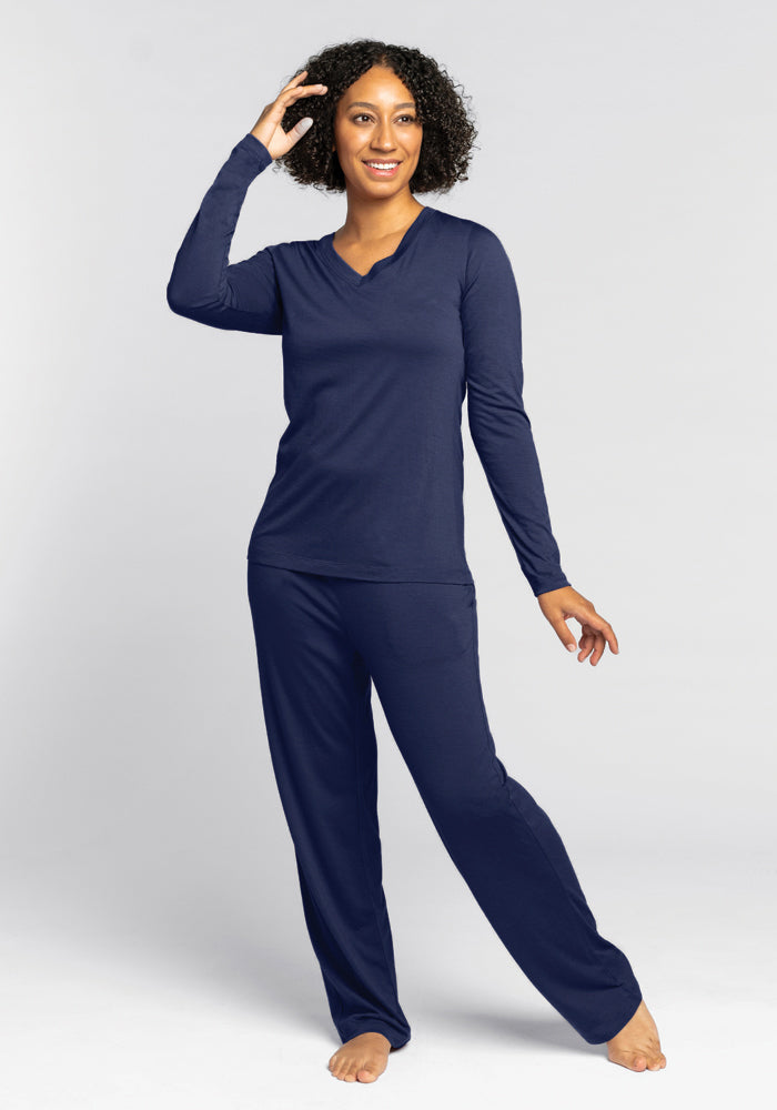 A person with curly hair stands smiling and posing in the cozy Lily V Neck Lounge Top - Starry Night by Woolx, known for its odor-resistant qualities. Against a plain white background, they are barefoot and casually touching their head with one hand.