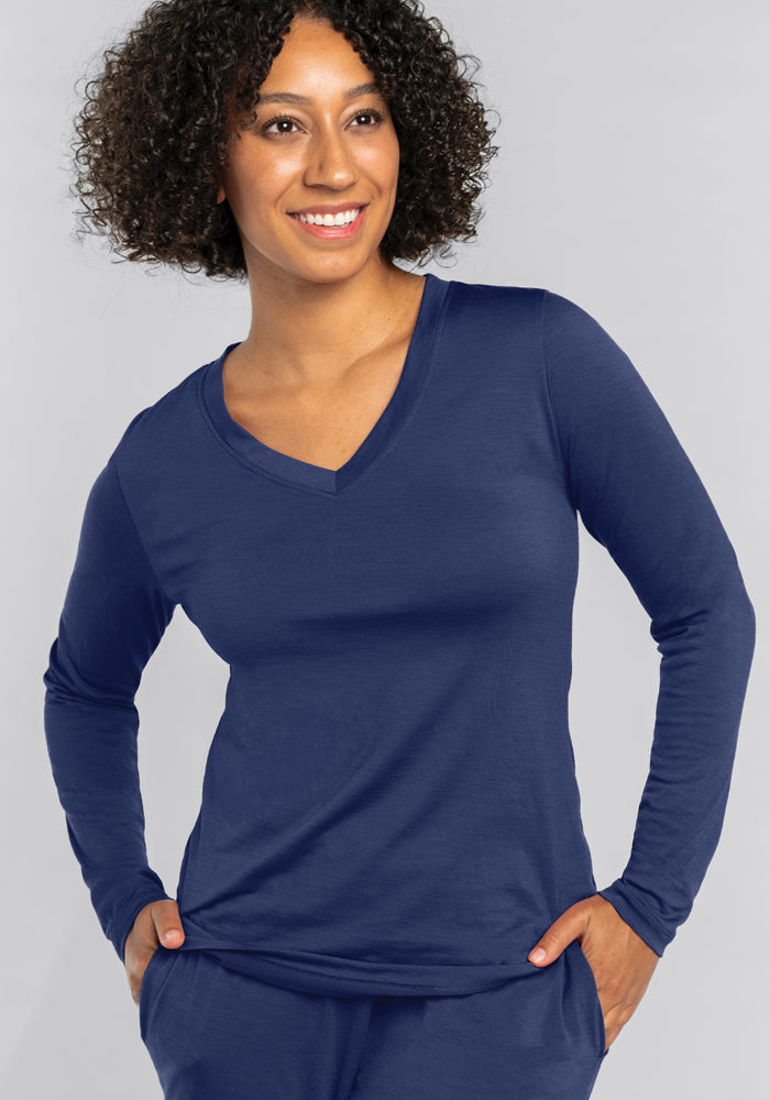 A person with curly hair is smiling while wearing a navy blue Lily V Neck Lounge Top in the Starry Night color, paired with matching pants. They are standing against a gray background with hands in their pockets, embodying the comfort provided by Woolx's Merino wool products.