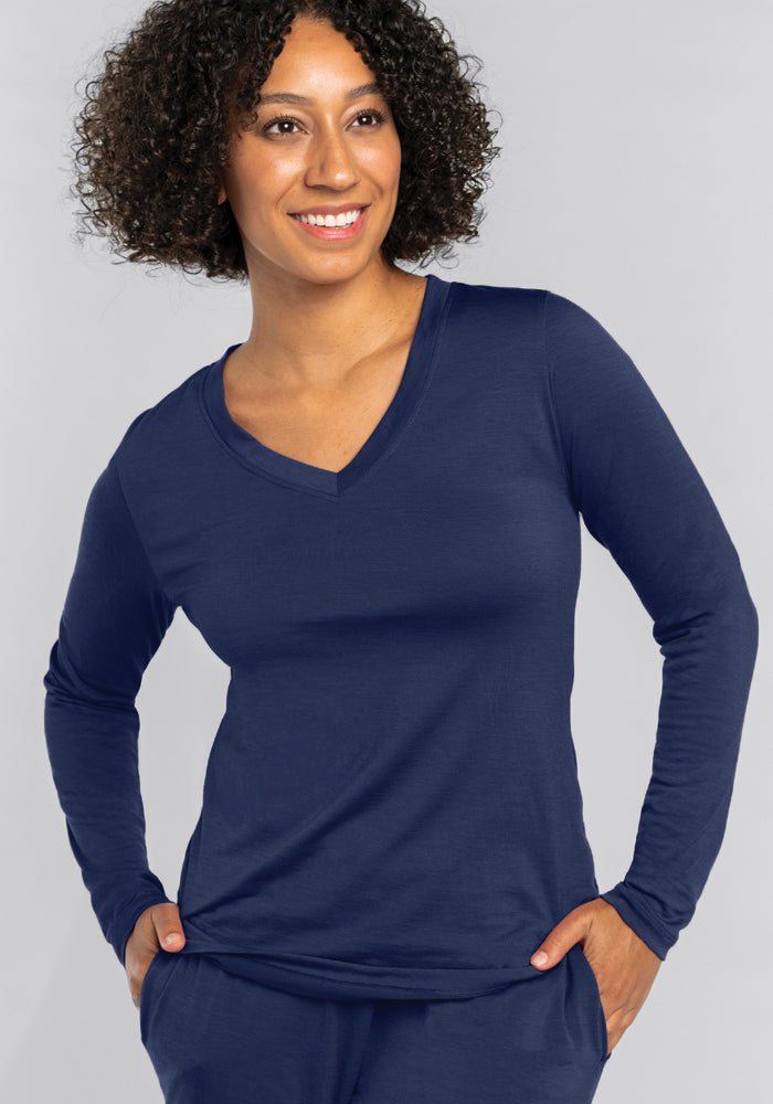 A person with curly hair smiles while wearing the Lily V Neck Lounge Top in Starry Night by Woolx, along with matching pants made from moisture-wicking Merino wool. They have their hands in their pockets and stand against a plain gray background.