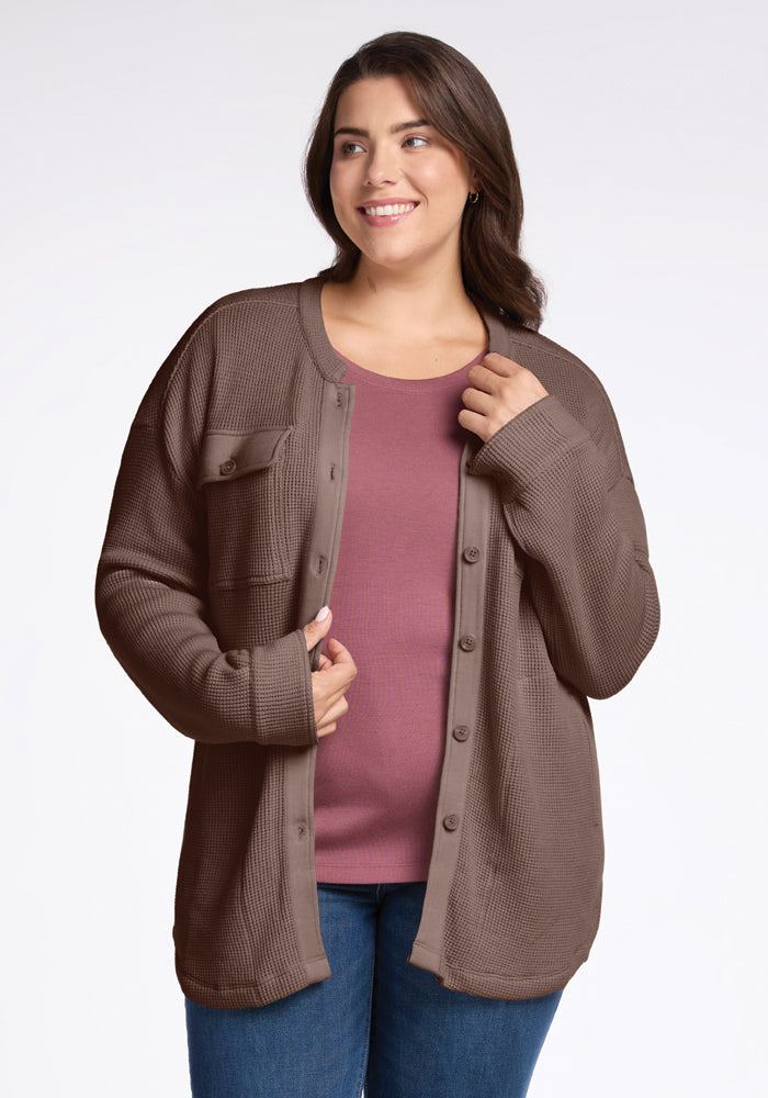 A woman smiles while looking to the side, exuding comfort in her Woolx Oaklynn Waffle Shacket in Latte. She pairs the shacket with a pink top and blue jeans, has long, dark hair, and stands against a plain white background. 