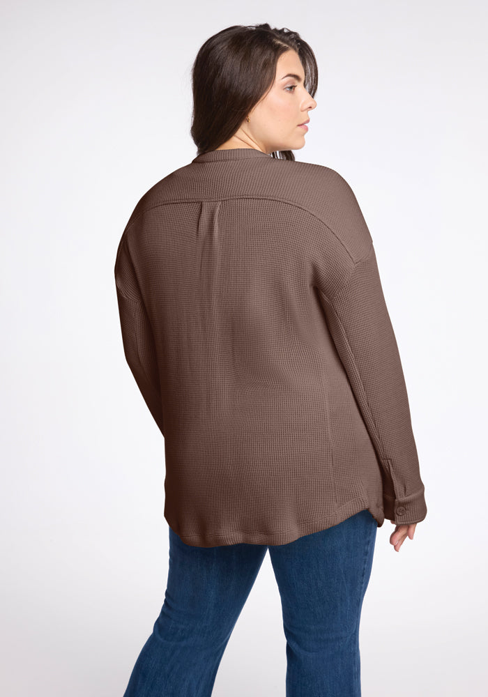 With long hair cascading down, an individual is turned away, displaying the cozy Oaklynn Waffle Shacket in Latte by Woolx over blue jeans. The backdrop is a simple, light-colored setting.