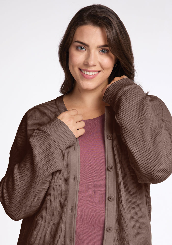 A person with long brown hair is smiling, wearing the Oaklynn Waffle Shacket in Latte by Woolx over a moisture-wicking mauve shirt. The plain, light-colored background enhances the cozy vibe.