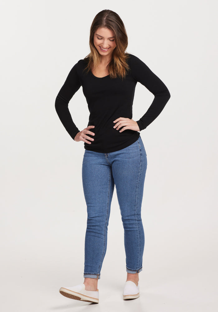 Wearing a Layla V Neck Top in black from Woolx, paired with blue jeans, a woman stands with her hands on her hips, smiling. Her light brown hair complements her white sneakers, ideal for an odor-resistant day out. The background is plain white.