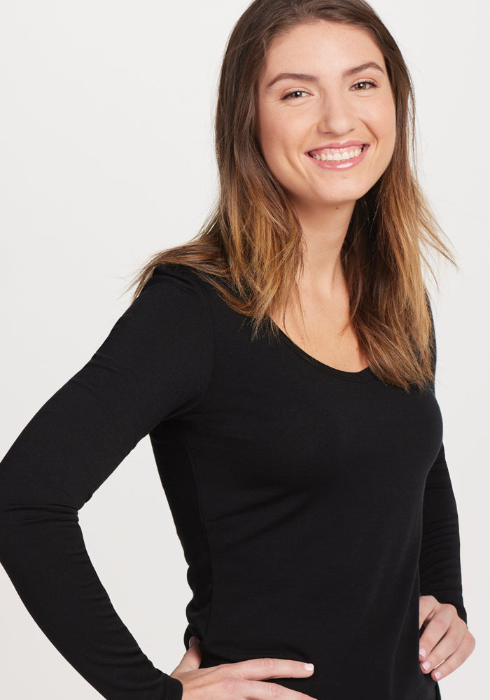 A person with a big smile and long brown hair stands against a plain white background, confidently placing their hands on their hips while wearing the Woolx Layla V Neck Top in black, crafted from moisture-wicking Merino Wool.