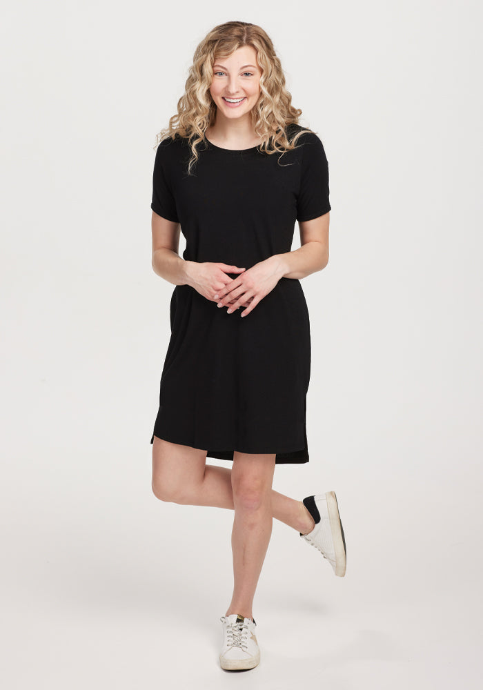 A person with curly hair smiles against a white background, wearing a Woolx Black Lexie Dress & Cover-up paired with white sneakers. Their hands are clasped in front, exuding effortless style and comfort. 