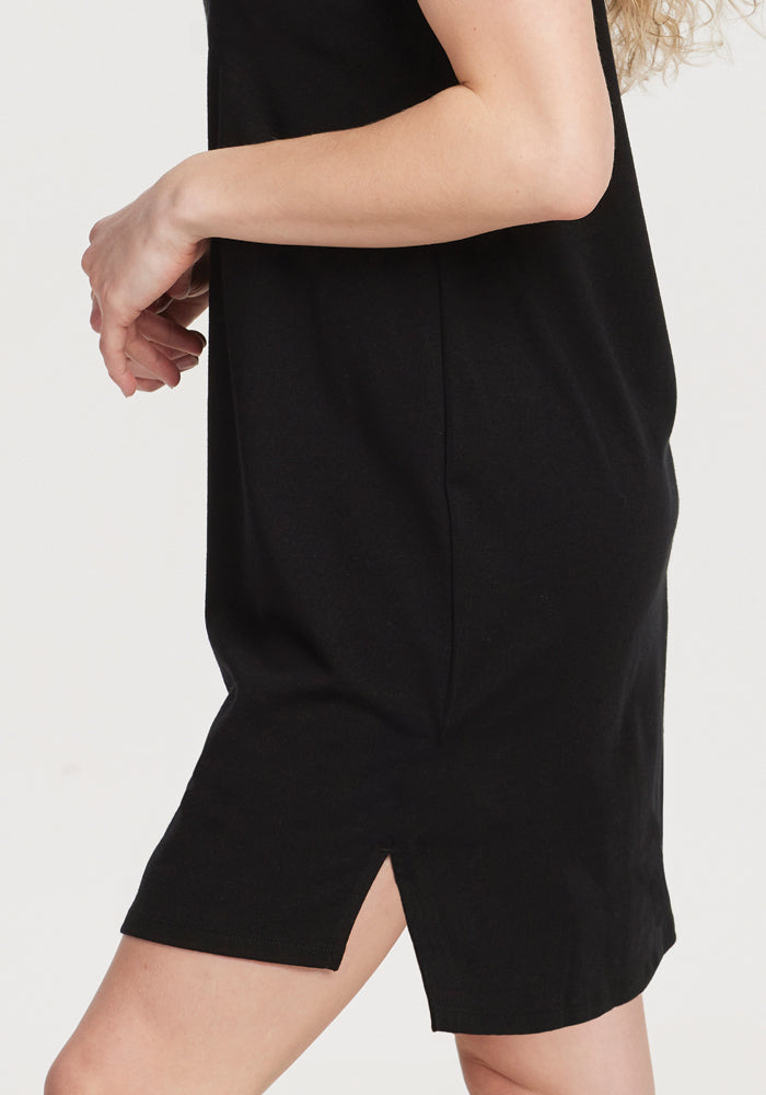 A person wearing the Woolx Lexie Dress & Cover-up in black is partially visible. The side view showcases its simple design, merino wool fabric, and a modest hemline slit, while slightly bent arms accentuate the dress's timeless elegance.