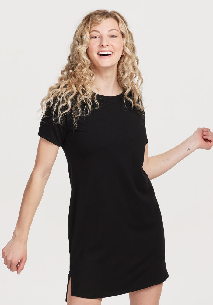 A person with curly blonde hair is smiling and wearing a Black Lexie Dress & Cover-up from Woolx, made of merino wool. They stand against a plain white background, looking cheerful and relaxed.