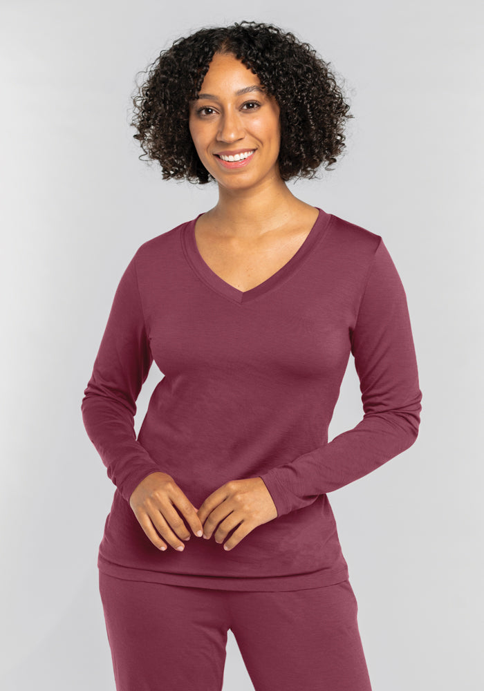 A person with curly hair smiles, wearing a moisture-wicking Woolx Lily V Neck Lounge Top in Wild Ginger. They stand against a light gray background, radiating style and comfort.