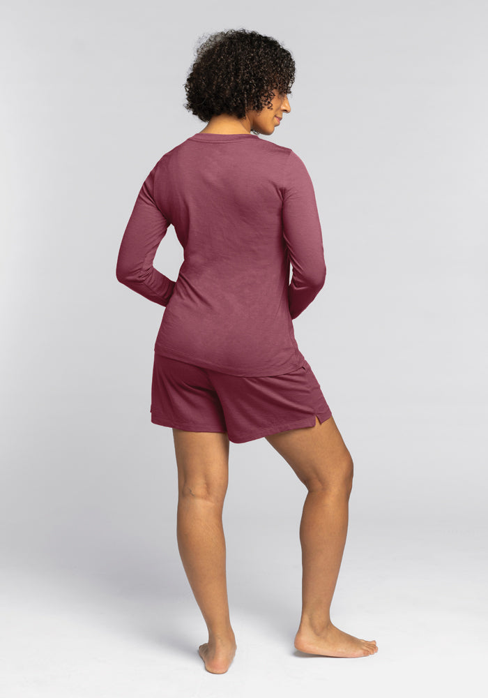 A person with curly hair is standing barefoot, facing away. They are wearing a matching sleepwear set, featuring a Lily V Neck Lounge Top and shorts in Wild Ginger Merino Wool by Woolx. The background is plain and light-colored.