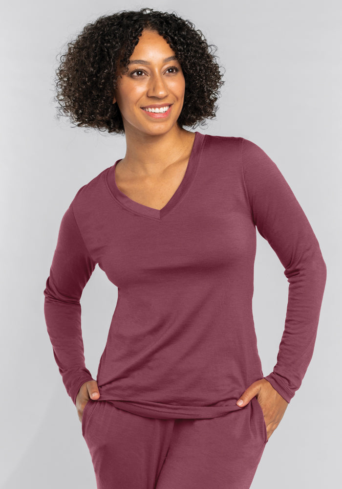 A person with curly hair smiles while wearing the Woolx Lily V Neck Lounge Top in Wild Ginger and matching pants. They are standing with one hand in a pocket against a light gray background, exuding comfort akin to cozy sleepwear.