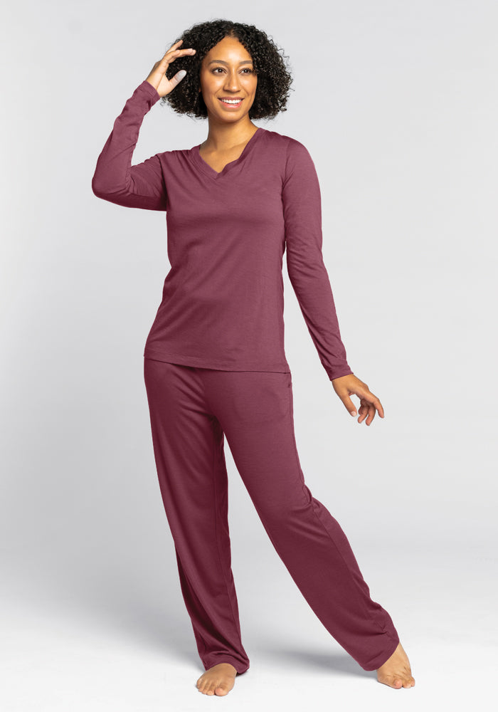 A woman with curly hair smiles as she wears a matching Woolx Lily V Neck Lounge Top in Wild Ginger. She stands barefoot against a plain gray background, one hand gently touching her head and the other resting by her side.