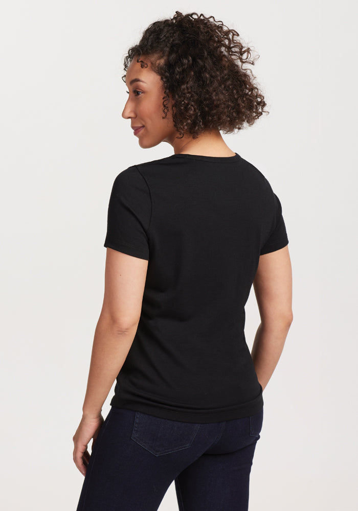 A person with curly hair is wearing Woolx's Liza Crew Neck Tee in black, crafted from soft merino wool, paired with dark jeans. They are standing and facing away from the camera against a plain white background. The moisture-wicking fabric ensures all-day comfort.