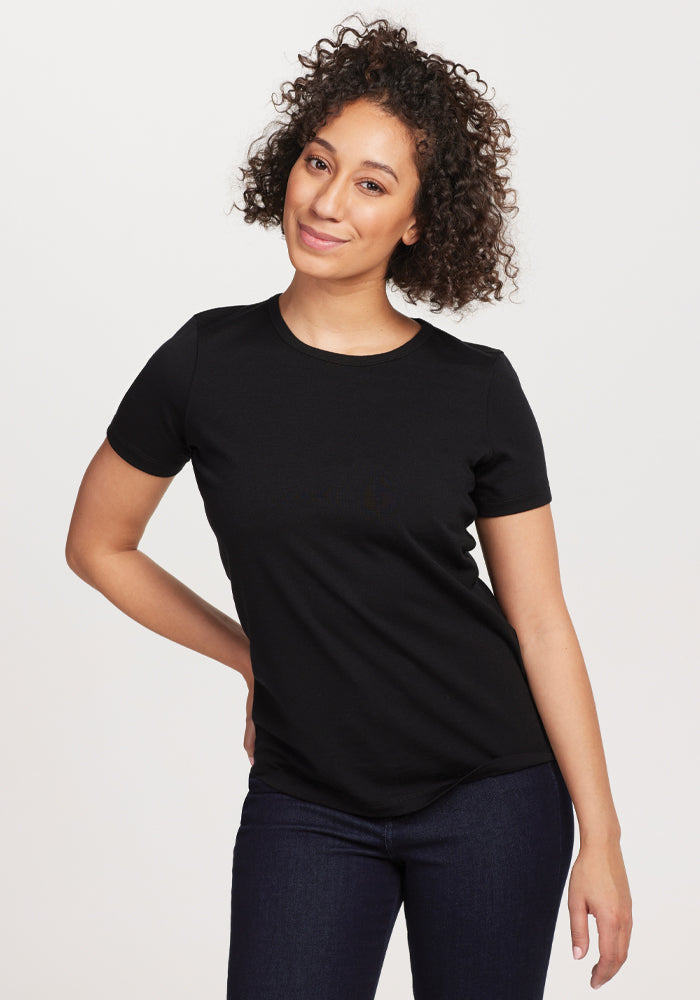 A person with curly hair stands smiling against a plain background, wearing the Woolx Liza Crew Neck Tee in Black made from moisture-wicking fabric, along with dark jeans. One hand rests on their hip while the other hangs by their side. 