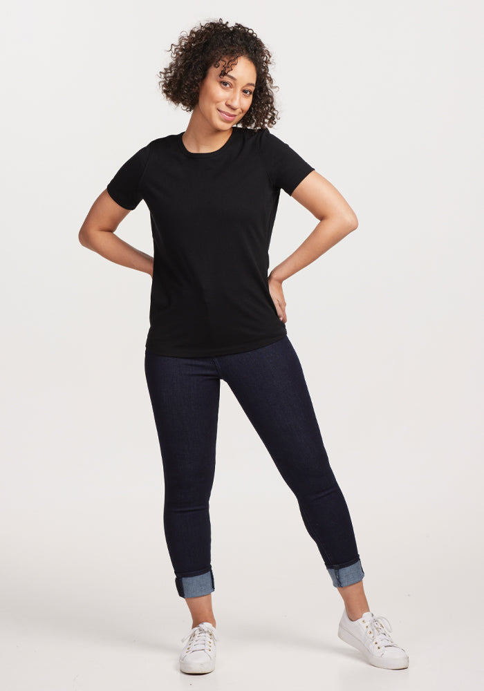 A person with curly hair stands confidently against a plain background, wearing the Woolx Liza Crew Neck Tee in black, crafted from moisture-wicking fabric, paired with dark jeans and white sneakers. Their hands rest on their hips as they smile slightly.