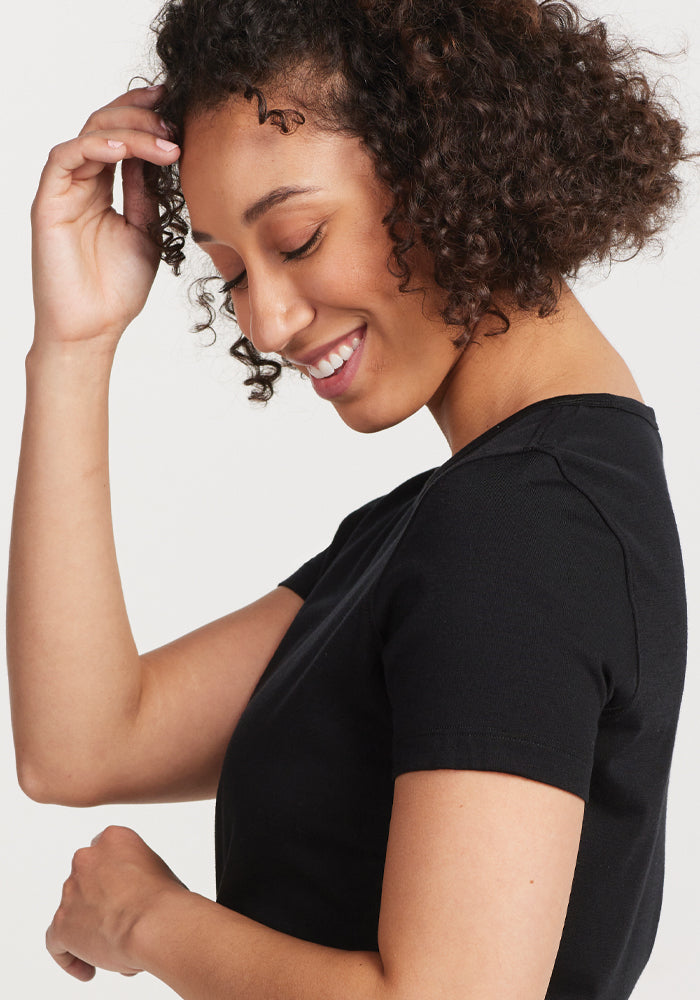 A person with curly hair smiles while touching their hair, wearing a Woolx Liza Crew Neck Tee in black, crafted from moisture-wicking fabric. They are looking down against a plain, light-colored background.