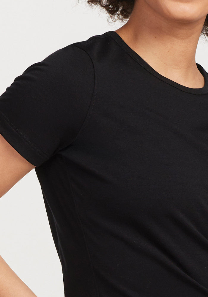 A close-up reveals a person in the Woolx Liza Crew Neck Tee - Black, crafted from soft merino wool. The upper torso and part of the head are visible, highlighting the short sleeves and round neckline. The curly-haired individual stands against a plain, light background.