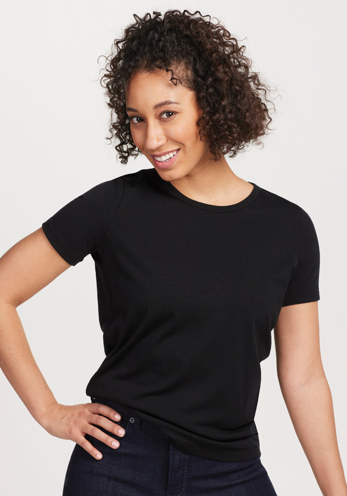 A person with curly hair wearing the Woolx Liza Crew Neck Tee in black, crafted from moisture-wicking merino wool, smiles and poses with hands on hips against a plain background.