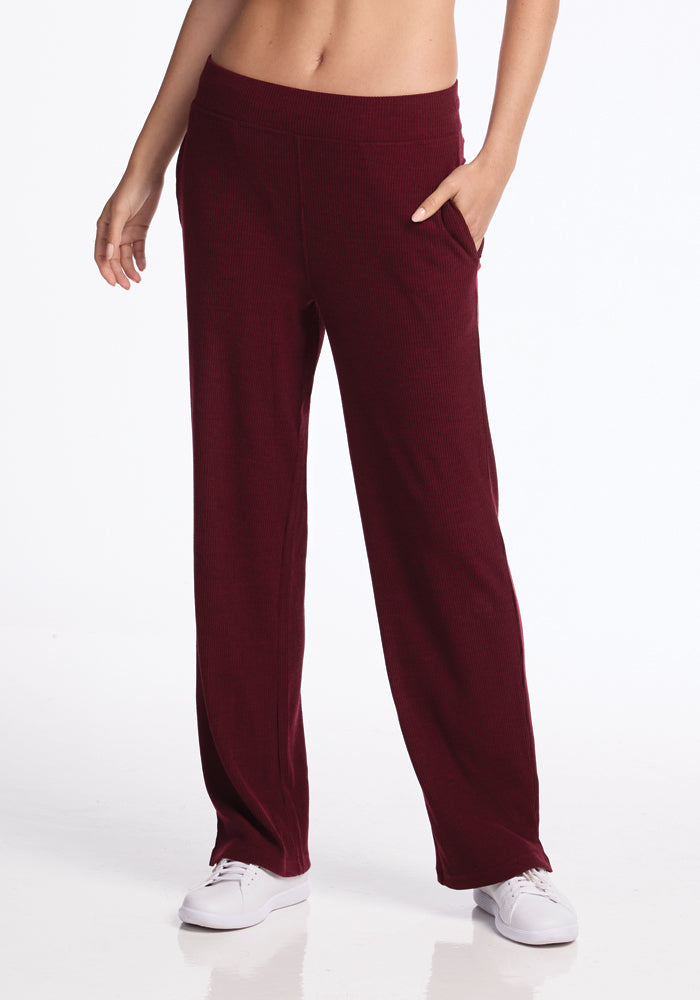 A person wearing Woolx's Luca Ribbed Pants in Cranberry Melange with their left hand in a pocket. The pants feature a relaxed, wide-leg fit and are paired with white sneakers against a plain white background. 