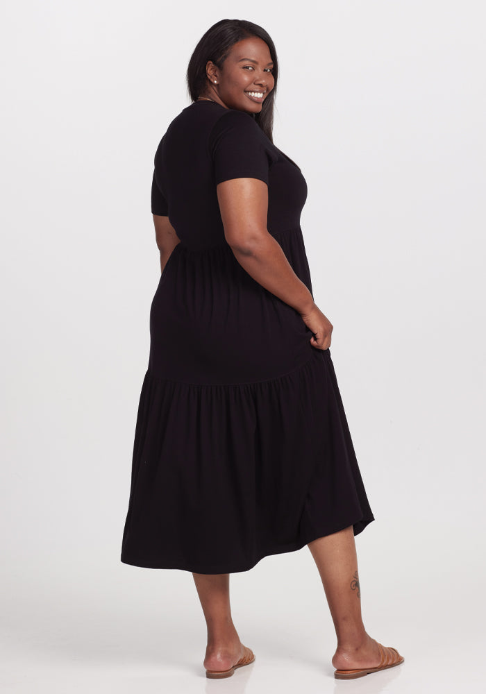 A person with long hair stands barefoot against a neutral background, smiling while looking over their shoulder in a Woolx Lucia Dress - Black, featuring tiered ruffles.