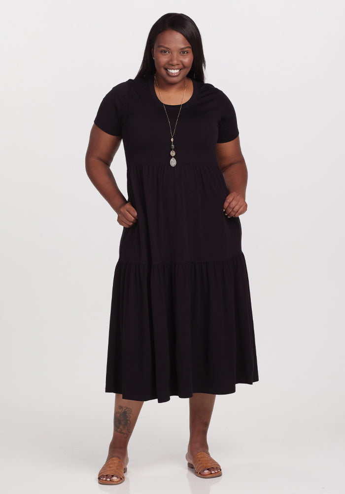 A woman smiles while posing in a black, short-sleeved Woolx Lucia Dress made of temperature-regulating merino wool. She accessorizes with a long necklace and sandals, her hands in the dress pockets, against a white backdrop. 