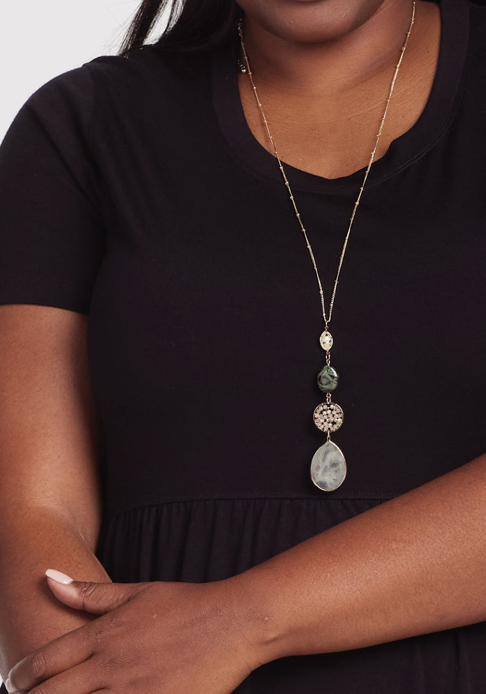 A person in a Woolx Lucia Dress - Black, paired with a long necklace having multiple pendants and a teardrop-shaped stone, crosses their arms comfortably.