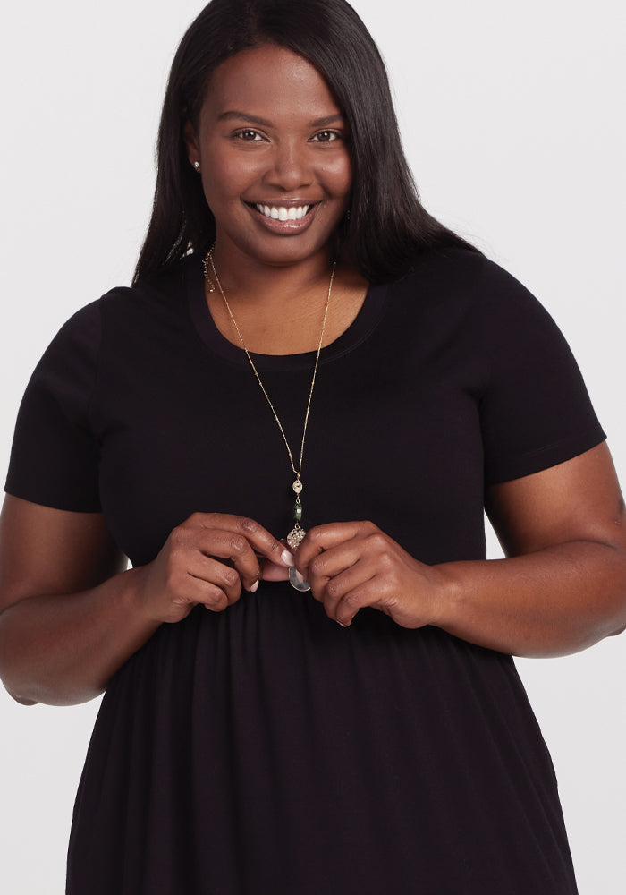 A person smiles warmly, wearing a Woolx Lucia Dress - Black made of temperature-regulating merino wool, against a plain white background. Their hands gently hold the pendant of a long necklace.