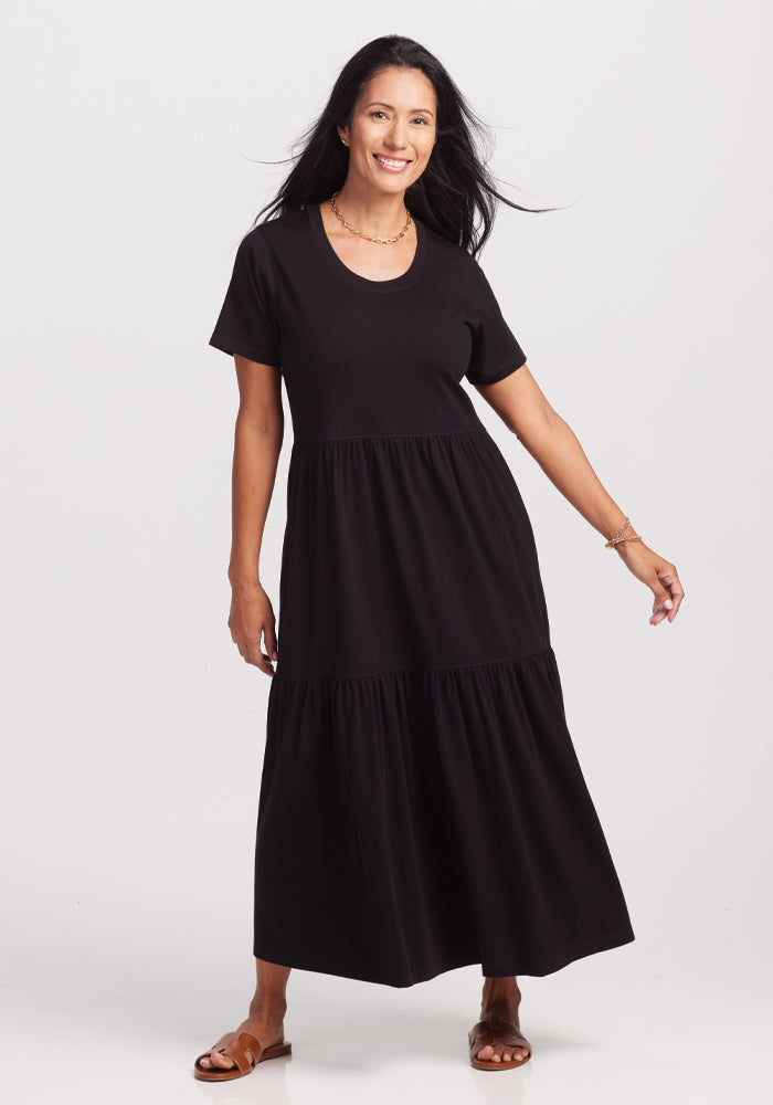 A woman with long black hair smiles in a black short-sleeve, tiered Lucia Dress by Woolx, crafted from temperature-regulating merino wool. She's on a white background, accessorized with a necklace and bracelet, wearing brown sandals. 