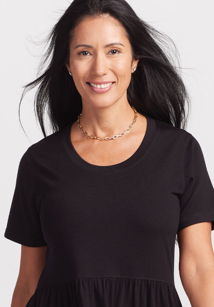 A woman with long dark hair smiles in a black Woolx Lucia Dress, adorned with a gold chain necklace, against a light backdrop, exuding effortless charm.