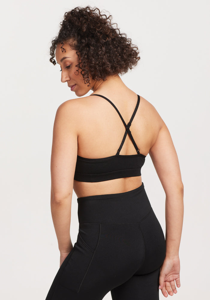 A person with curly hair wears the Luna Bralette - Black from Woolx, featuring moisture-wicking material and crisscross straps, paired with matching athletic leggings. They stand against a plain white background, looking over their shoulder.