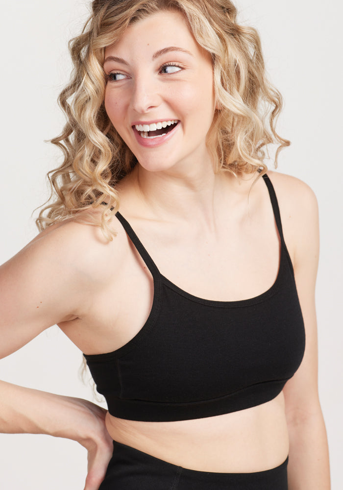 A person with curly blonde hair, smiling and looking to the side, is wearing the Woolx Luna Bralette in black. The plain, light-colored background perfectly highlights their cheerful expression. 