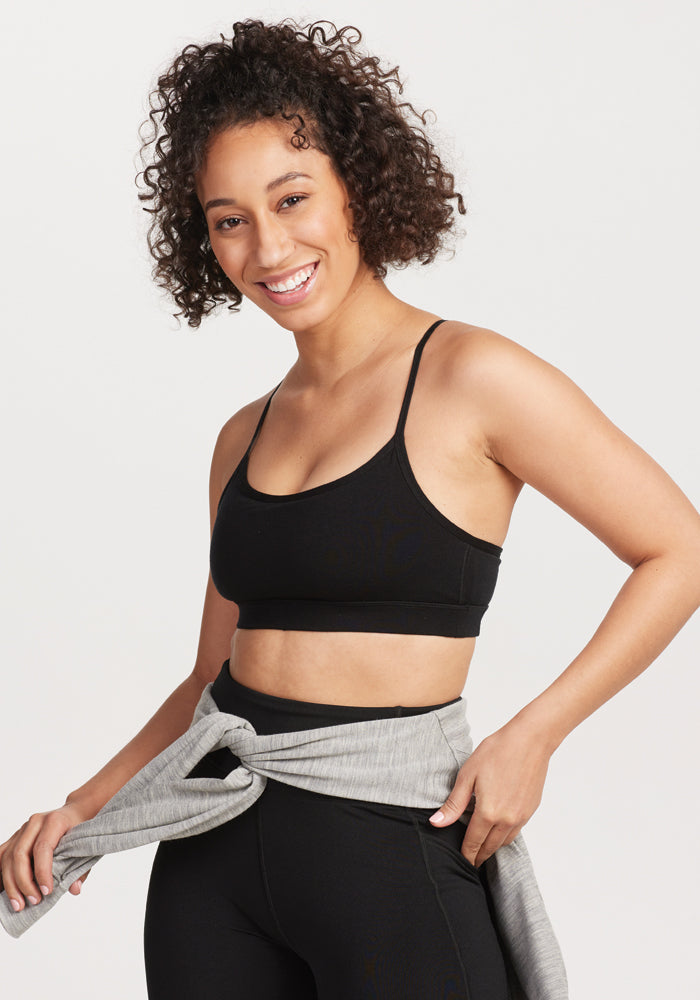 A person with curly hair smiles while wearing a black Luna Bralette by Woolx and moisture-wicking black leggings. They have a gray sweatshirt tied around their waist and stand against a plain background. 