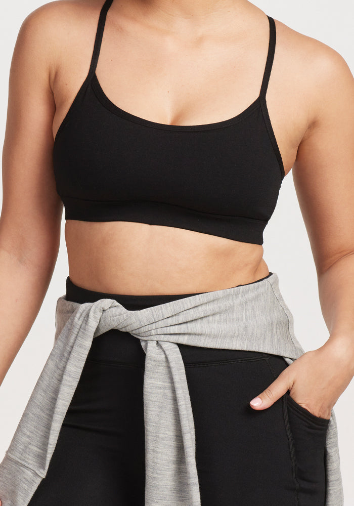 Wearing a Woolx Luna Bralette - Black and leggings, the individual has a light gray merino wool sweater tied around their waist. One hand rests in their pocket against a plain white backdrop.