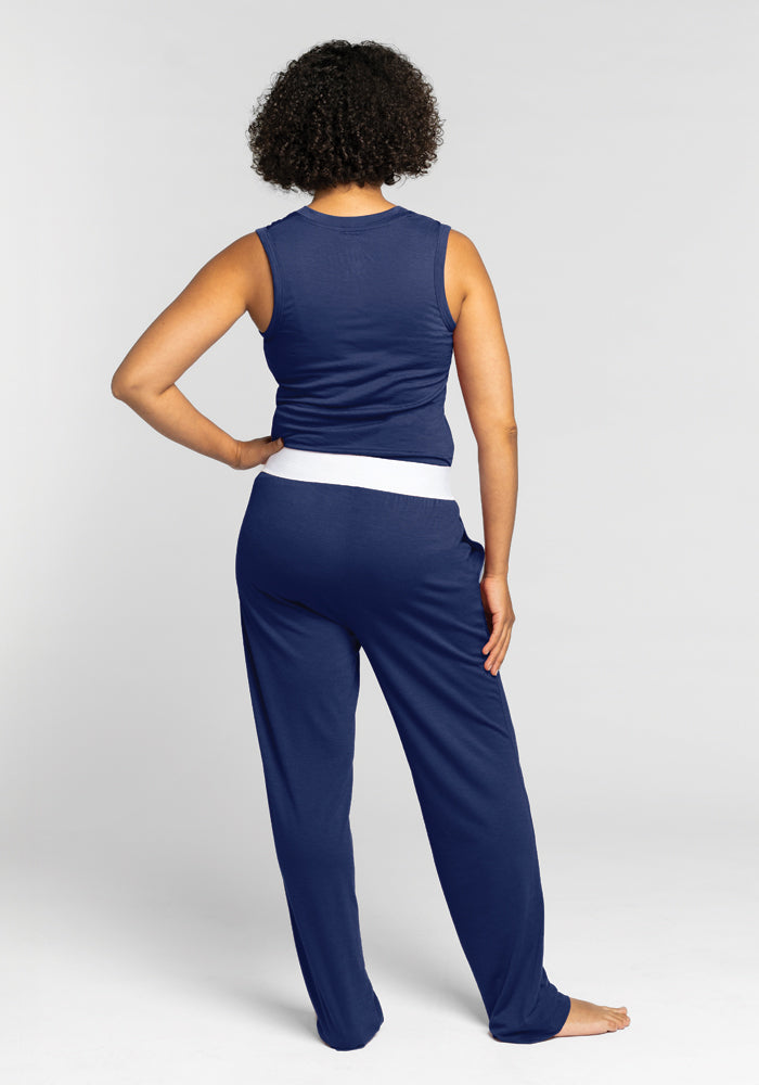 A person with short curly hair stands facing away, wearing a sleeveless navy top and Woolx's Maya Lounge Pants in Starry Night, featuring a comfortable white waistband, against a plain gray background.