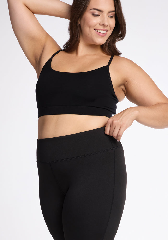 A woman smiling and posing in a black Woolx athletic outfit, featuring the Marissa Pants - Black as high-waisted leggings and a coordinating sports bra. She has one hand on her hip and the other raised behind her head against a plain white background.