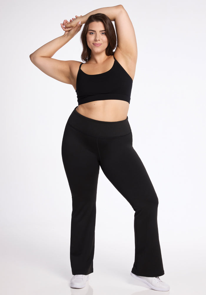 A person in a black sports bra and Woolx's Marissa Pants in black strikes a confident pose with arms raised. They are wearing white sneakers and have a relaxed expression against a plain white background.