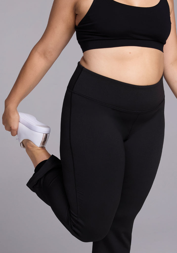 A person dressed in a black sports bra and Woolx's Marissa Pants stretches by holding their right foot behind them. They are wearing white sneakers, balancing on one leg against a light gray background.