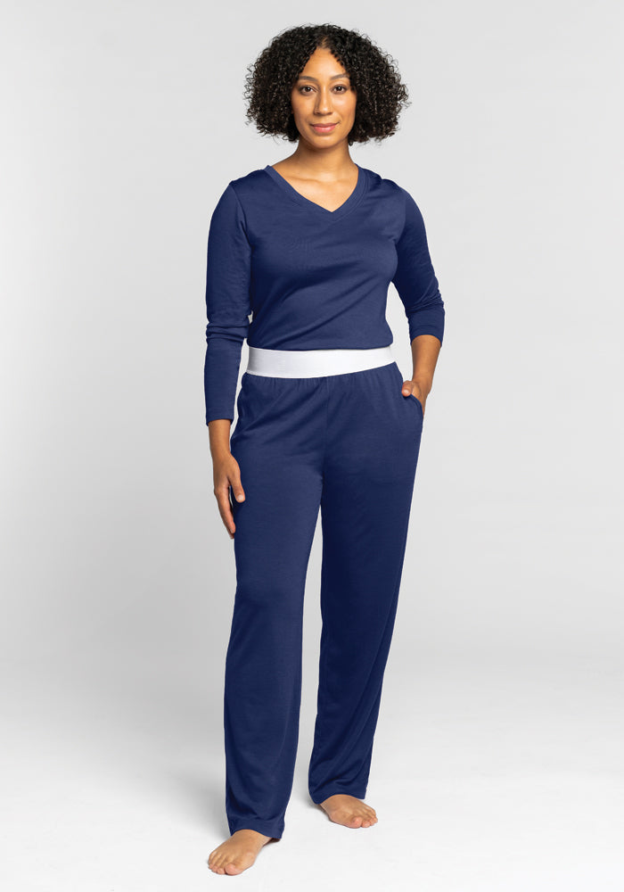 A person stands barefoot against a plain grey background, wearing a blue long-sleeve top and matching Woolx Maya Lounge Pants in Starry Night with a contrasting white waistband. Crafted from soft Merino wool, they have curly hair and look directly at the camera, with one hand tucked in a pocket.