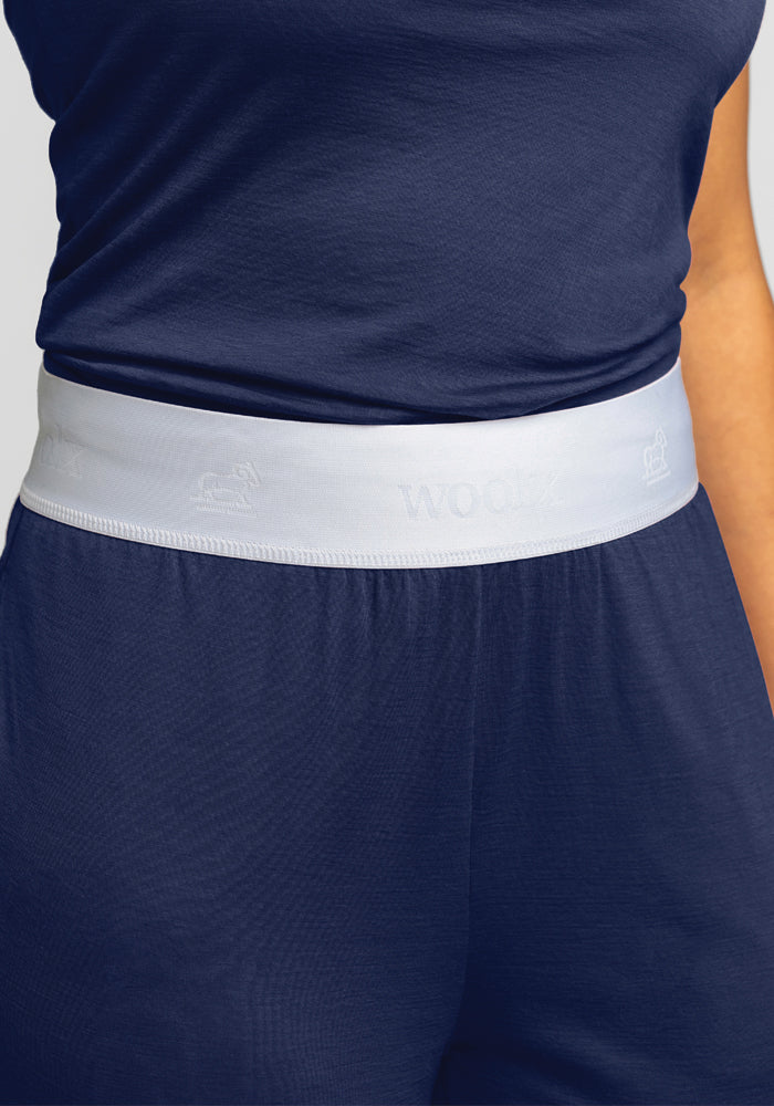 A close-up showcases a person wearing the Maya Lounge Pants - Starry Night by Woolx, which feature a light gray, comfortable waistband. The dark blue pajama pants are crafted from soft Merino wool, with the waistband subtly embossed with text and symbols. A plain background enhances the elegant simplicity of the image.